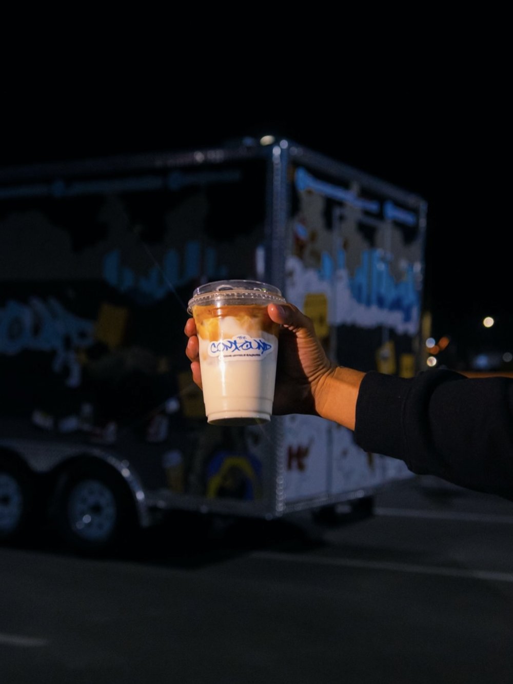 A hand holding a The Compound Coffee iced drink in front of the truck